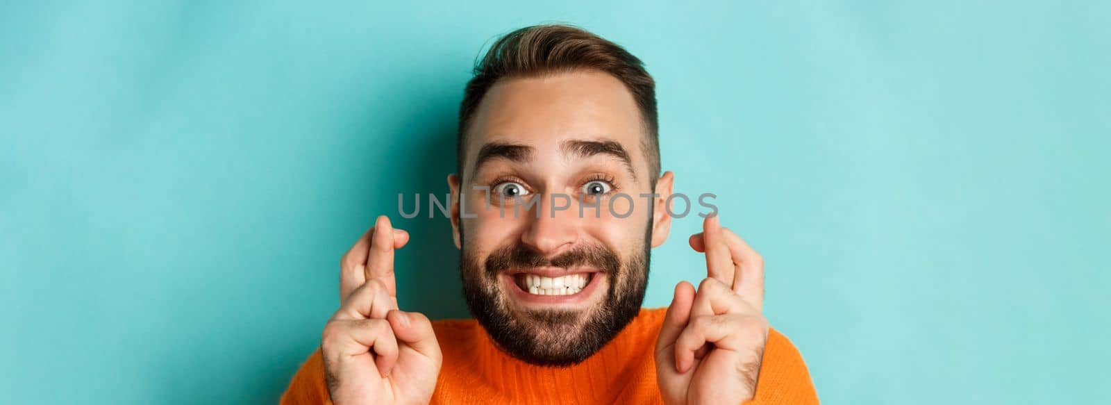 Headshot of hopeful bearded man making a wish, smiling and holding fingers crossed for good luck, standing over light blue background.