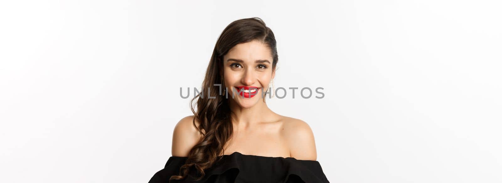 Close-up of beautiful woman dressed for party in black dress, wearing makeup and red lipstick, smiling happy at camera, white background.