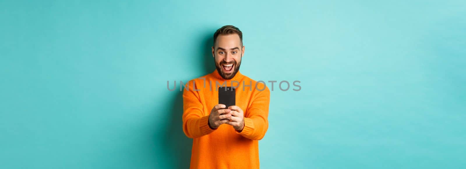 Image of man reading awesome message on mobile phone, looking at smartphone screen with amazement, standing over light blue background.