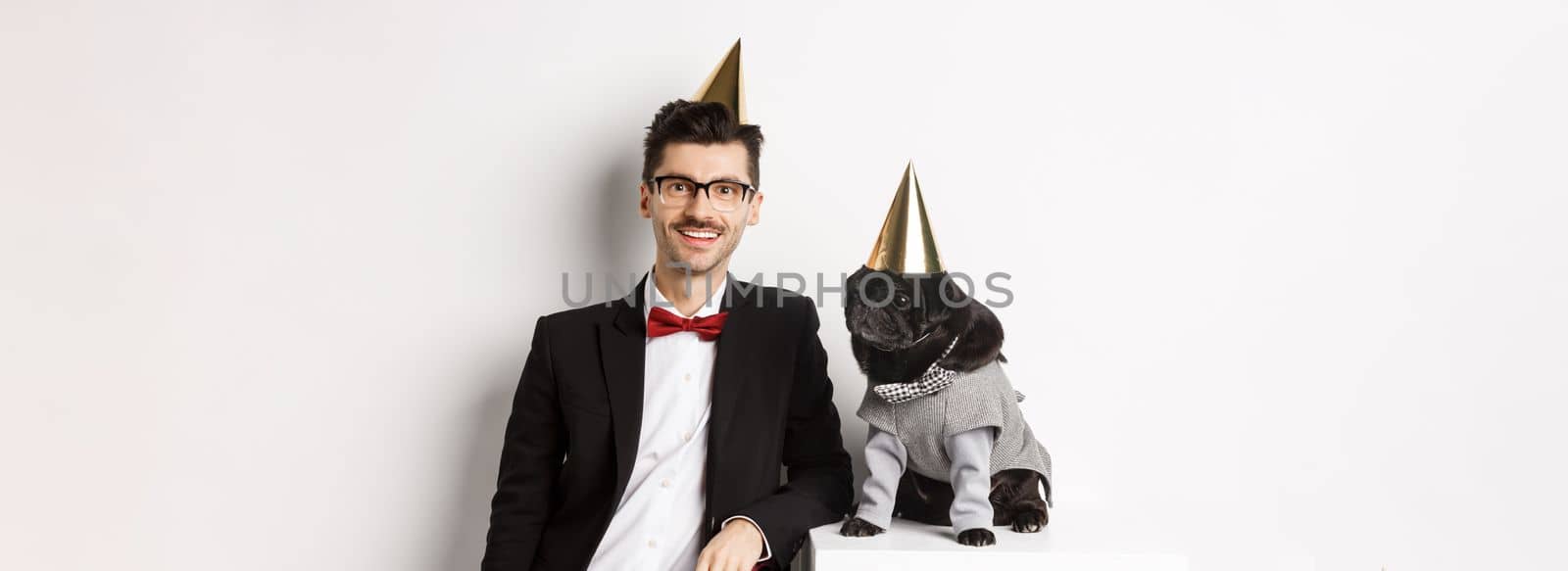 Image of handsome young man celebrating birthday with cute black pug in party costume and cone on head, standing over white background by Benzoix