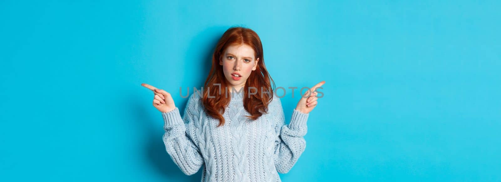 Confused redhead girl in sweater pointing fingers sideways, staring at camera doubtful, standing against blue background by Benzoix