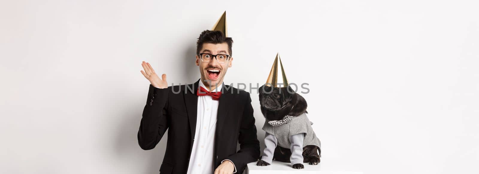 Happy young man and cute black dog wearing party cones, celebrating birthday, guy friendly saying hello and waving hand, standing over white background.