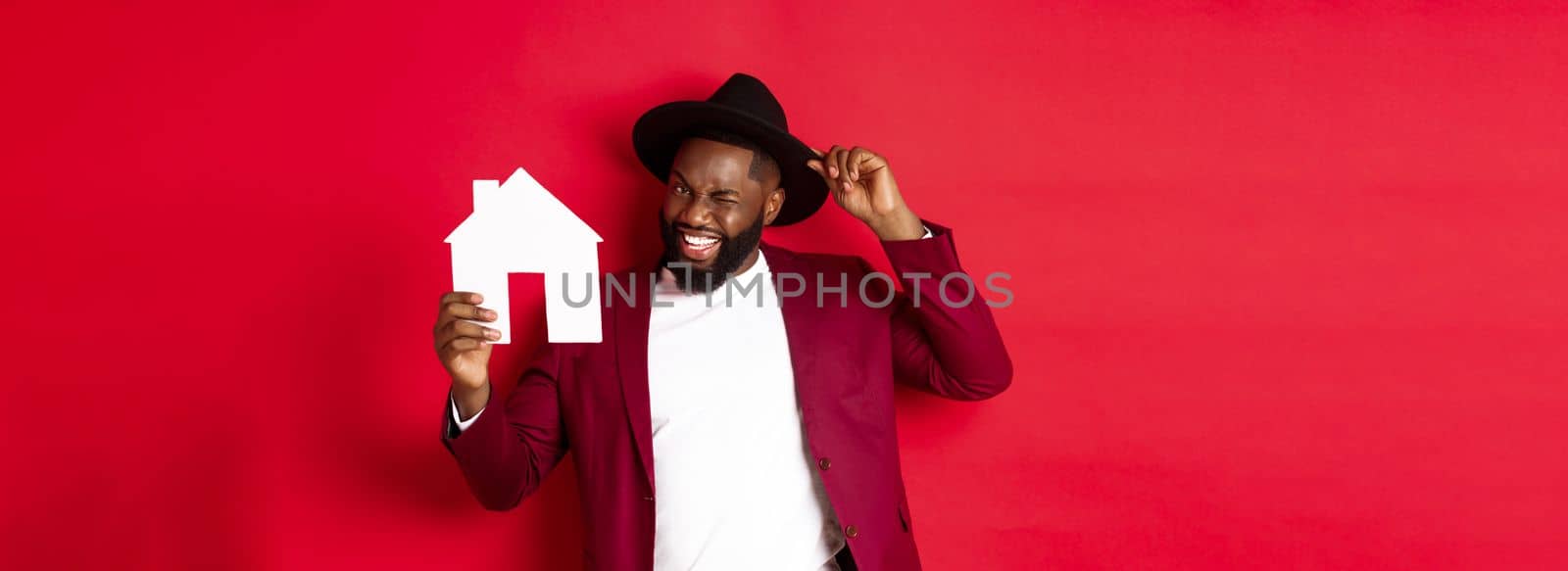 Real estate. Cheerful Black man showing paper house and smiling, recommending broker, standing over red background.