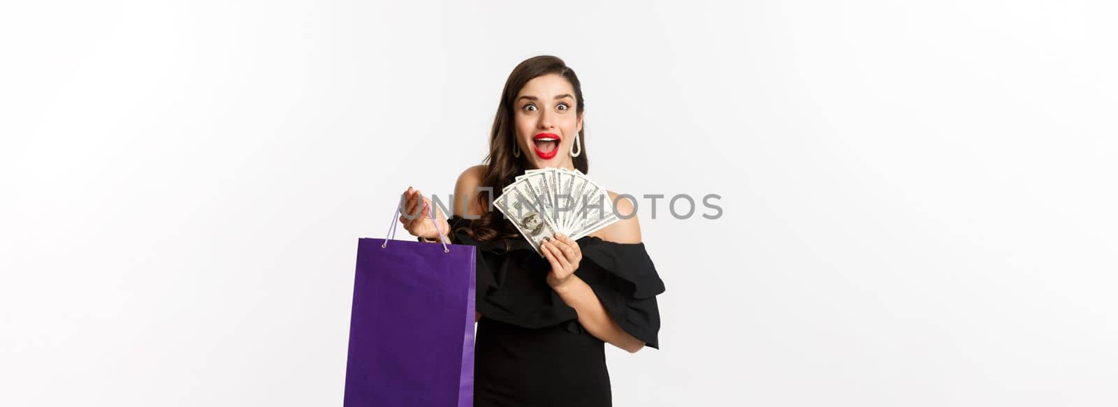 Image of excited beautiful woman in black dress going shopping, holding bag and dollars, standing over white background.