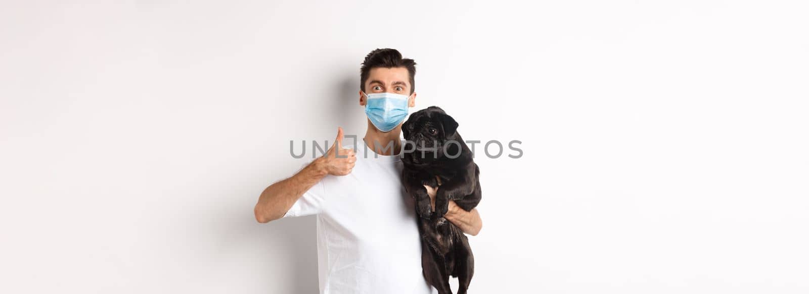 Covid-19, animals and quarantine concept. Young man in medical mask holding cute black pug dog, showing thumb up, like and approve, standing over white background by Benzoix