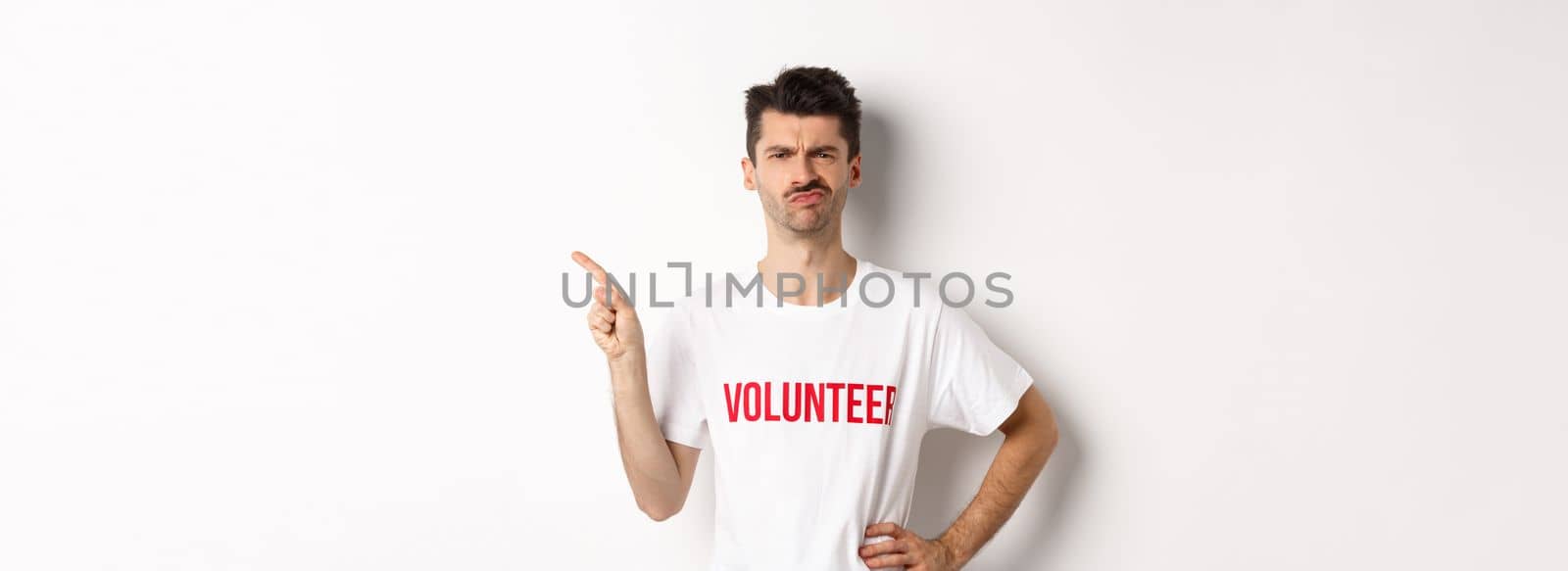 Skeptical and hesitant male volunteer in t-shirt grimacing doubtful, pointing finger left at promo offer, white background.