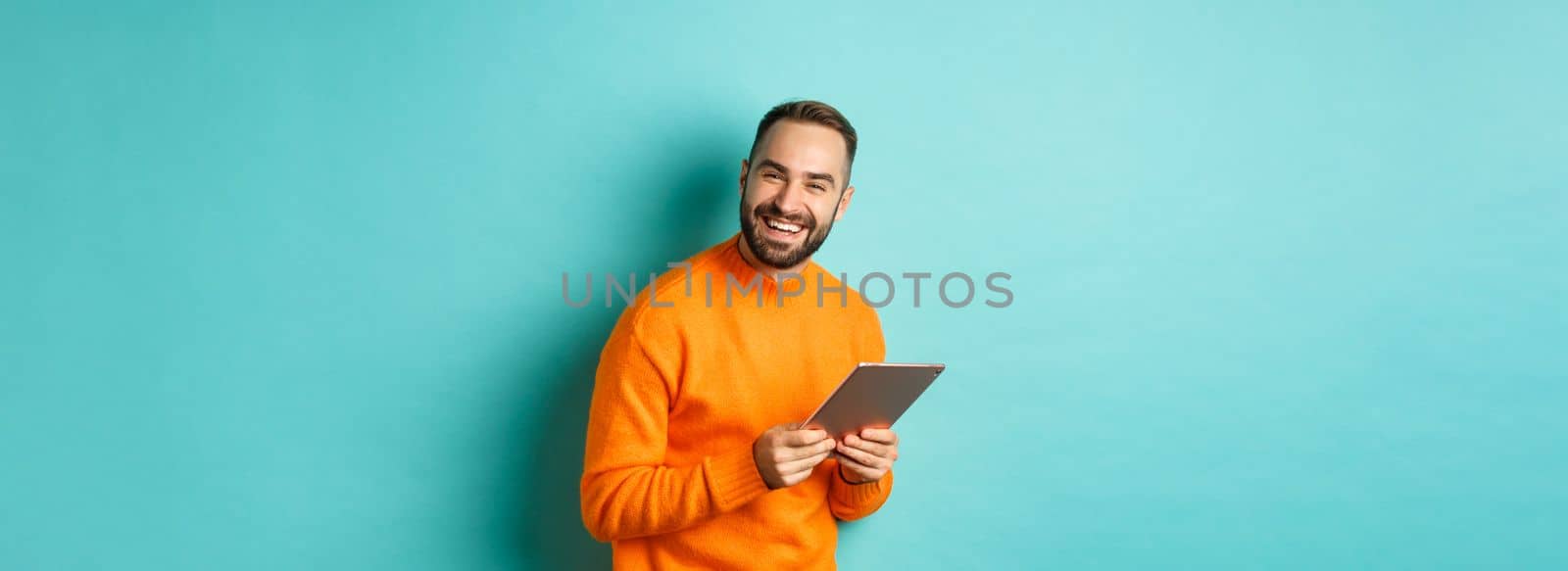 Handsome bearded man using digital tablet, laughing at camera, standing happy against light blue background by Benzoix