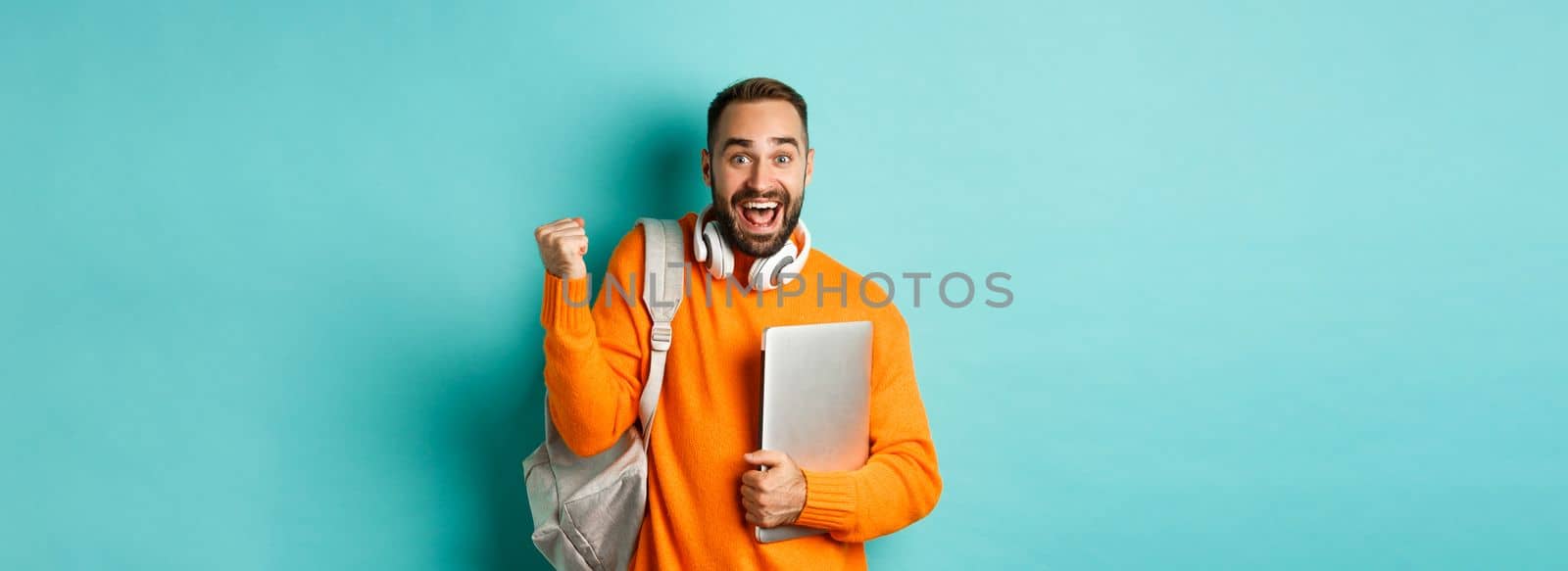 Happy man with backpack and headphones, holding laptop and smiling, cheering of win, triumphing over turquoise background.