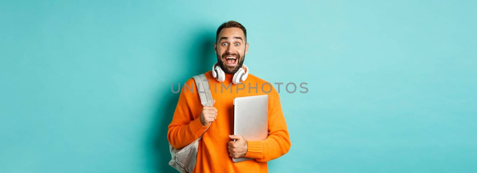 Happy man with backpack and headphones, holding laptop and smiling, looking excited, standing over turquoise background.