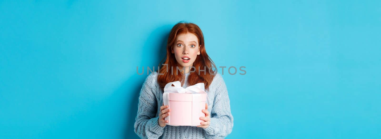 Surprised redhead girl receiving valentines gift, holding box with present and staring at camera amazed, wearing sweater, standing over blue background by Benzoix