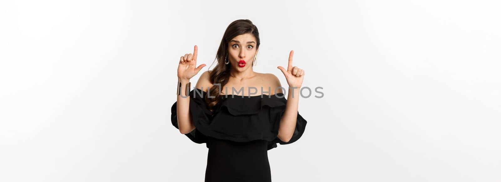Fashion and beauty. Surprised woman showing advertisement on top, pointing fingers up and saying wow amazed, standing over white background.
