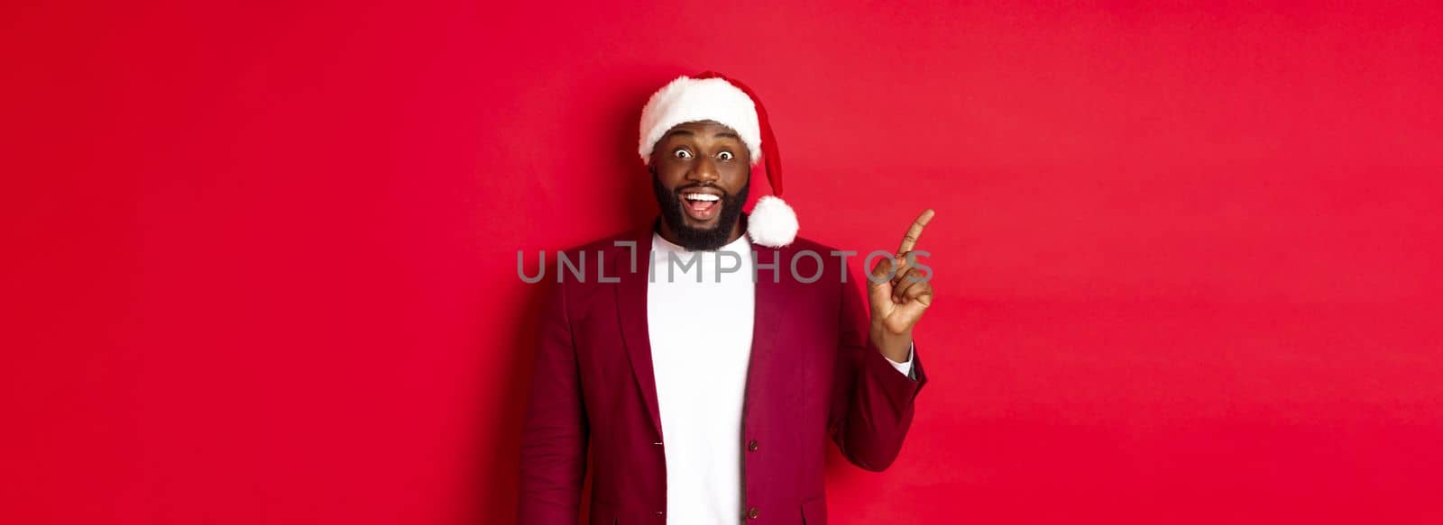 Christmas, party and holidays concept. Excited and amused Black man pointing finger left and smiling, showing logo, standing over red background.