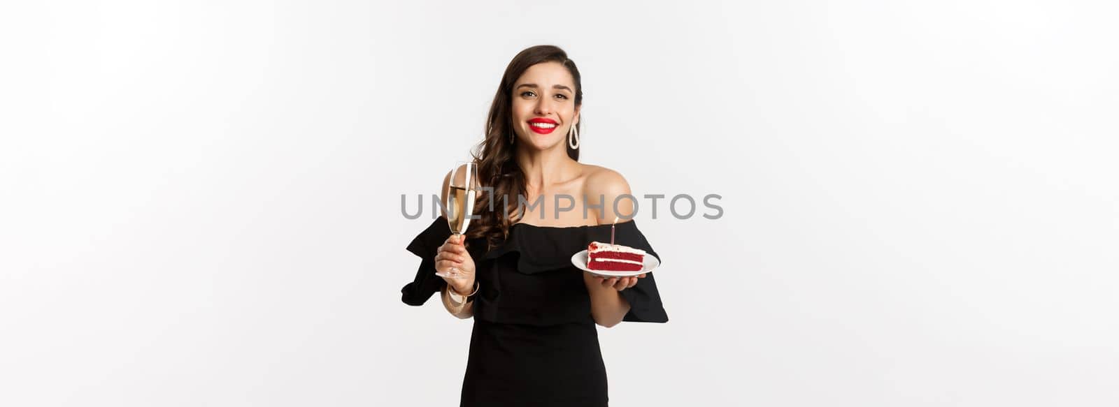 Celebration and party concept. Happy woman holding birthday cake and drinking champagne, smiling while standing in black dress against white background.