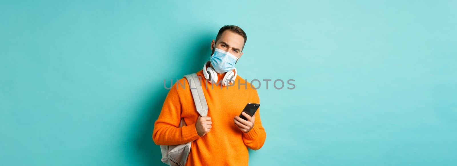 Skeptical and disappointed young man wearing face mask, holding backpack and mobile phone, frowning upset, standing over light blue background.