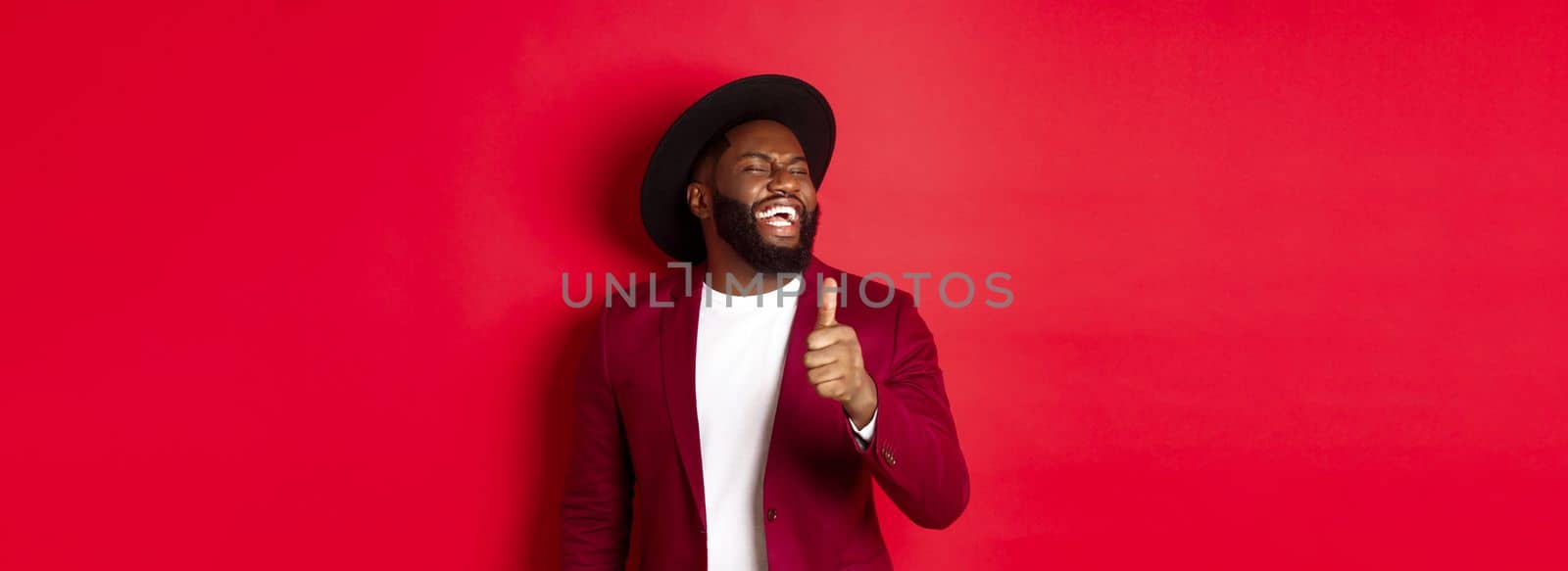 Handsome african american man having fun, showing thumb up and laughing from good joke, standing against red background by Benzoix