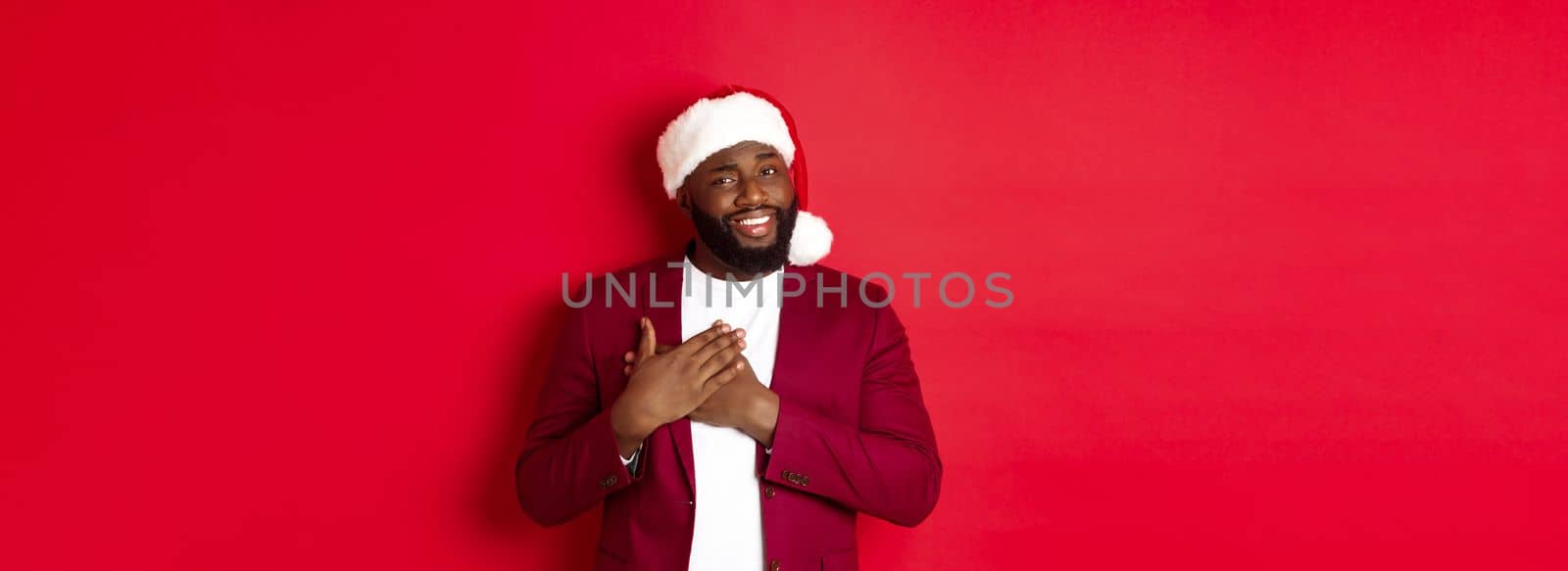 Christmas, party and holidays concept. Grateful african american man in santa hat saying thank you, holding hands on heart and smiling, feeling touched, standing against red background.