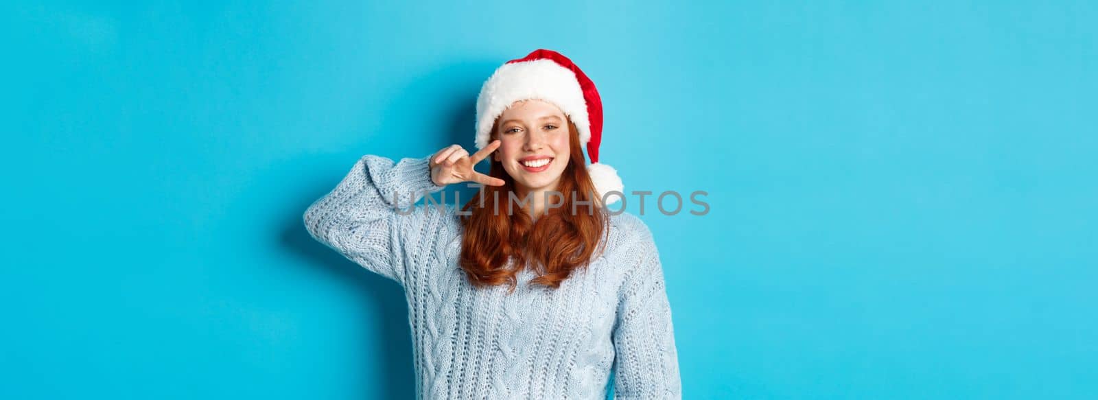 Winter holidays and Christmas Eve concept. Happy teenage girl with red hair, wearing santa hat, enjoying New Year, showing peace sign and smiling, standing over blue background.