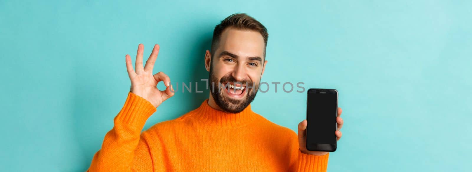 Photo of caucasian man showing mobile screen and okay sign, approve online store, smartphone app, standing satisfied over light blue background.