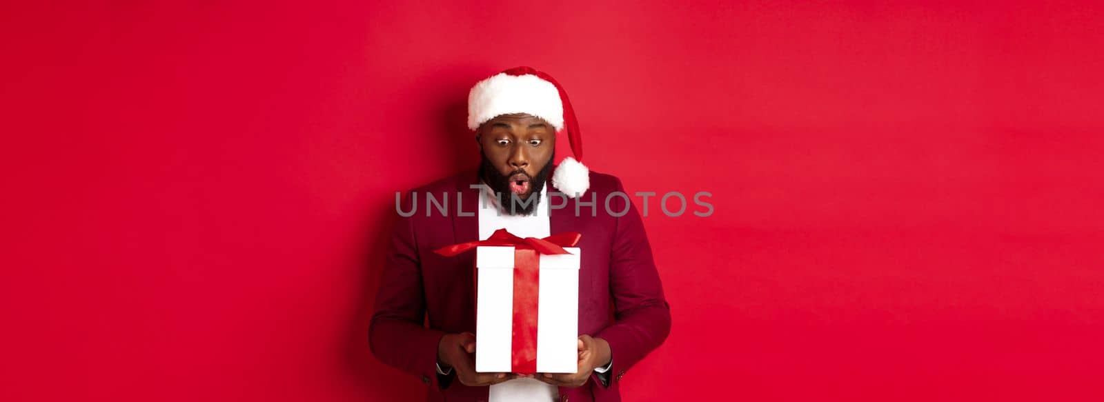Christmas, New Year and shopping concept. Surprised Black man staring at xmas present, saying wow amazed, receiving holiday gift, red background by Benzoix