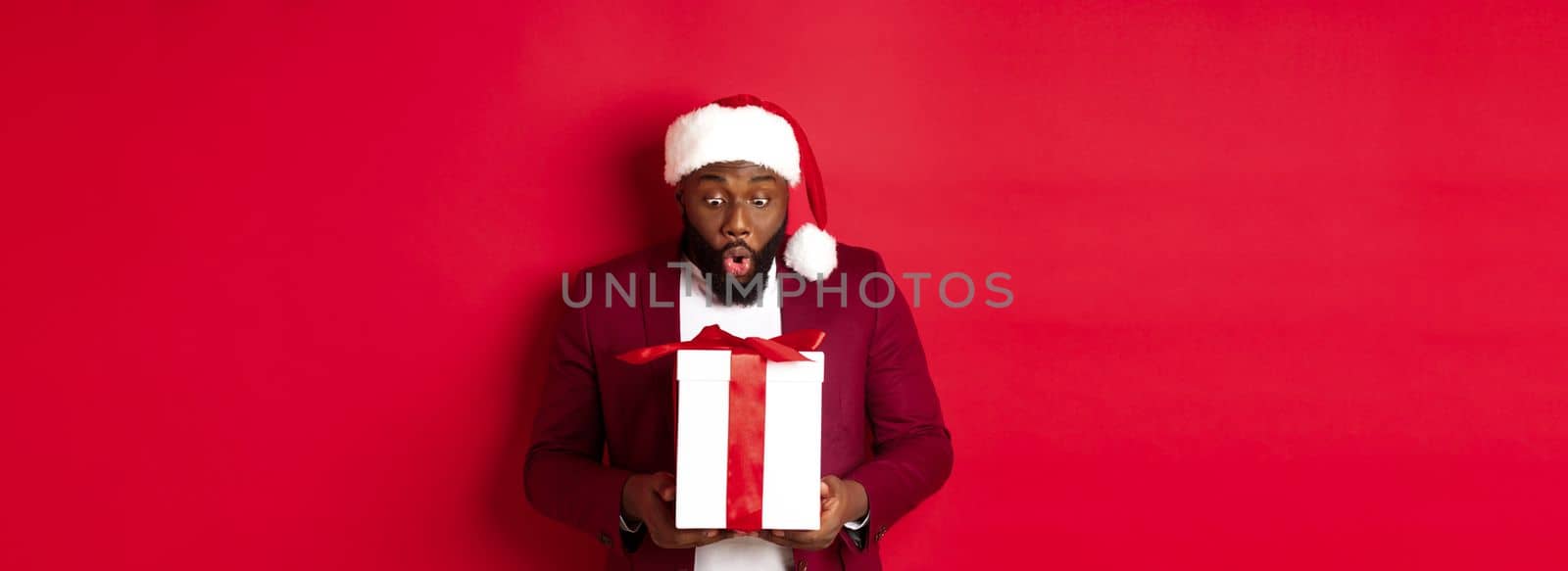 Christmas, New Year and shopping concept. Surprised Black man staring at xmas present, saying wow amazed, receiving holiday gift, red background.