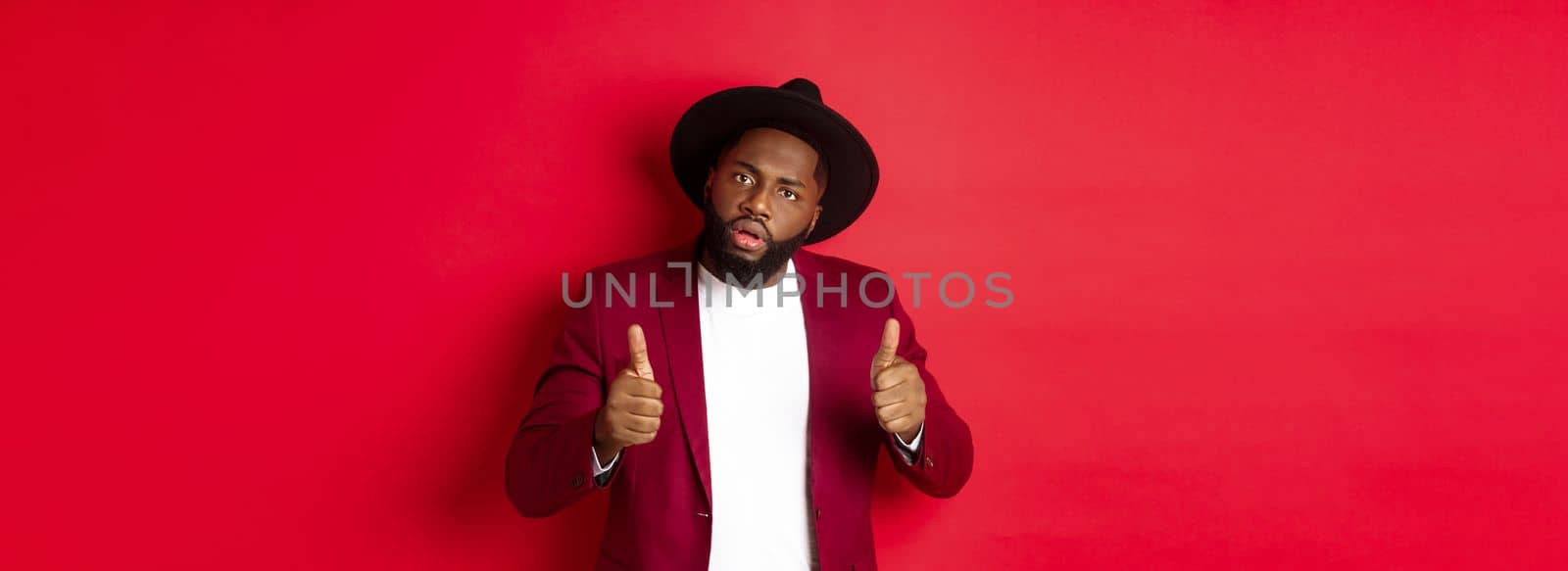 Impressed Black man saying good job, cheer up with thumbs-up, praise something, standing against red background. Copy space