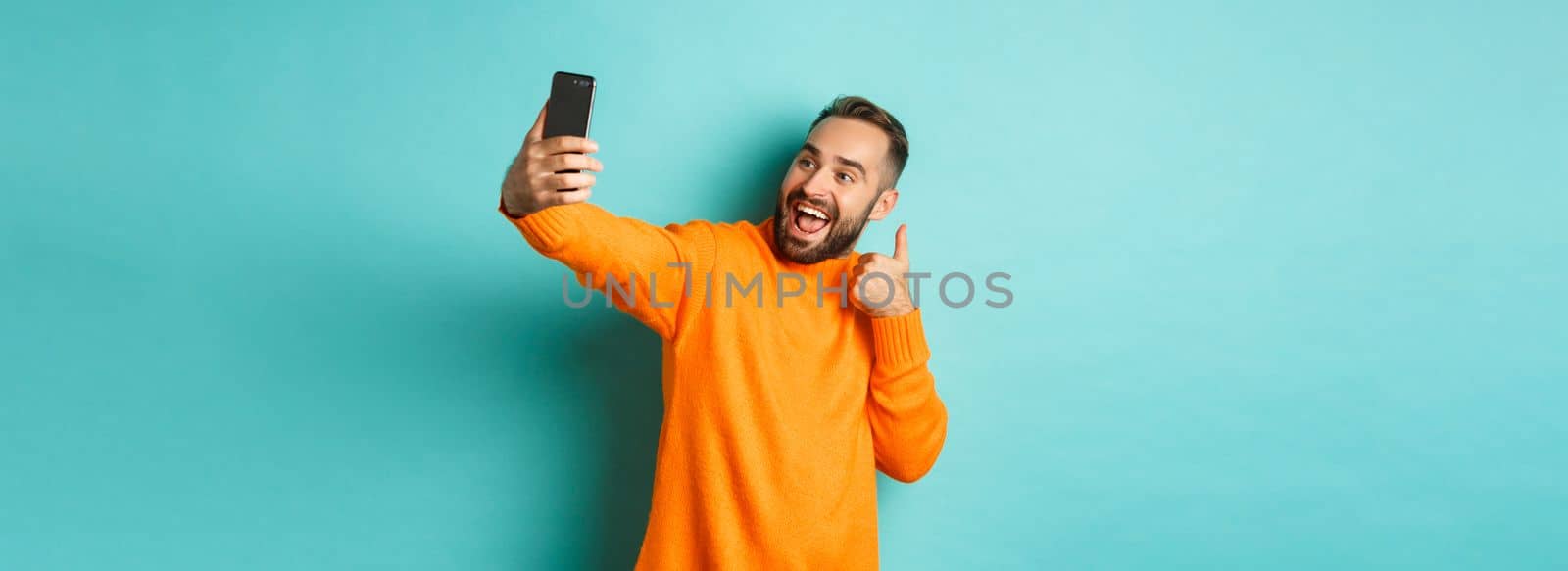 Handsome young man taking selfie on mobile phone, showing thumbs up at smartphone camera, recording vlogg, standing over light blue background.