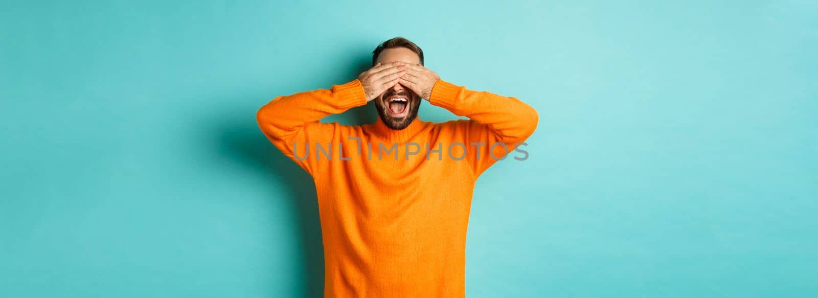 Happy bearded man waiting for surprise, shut ears and expeting gifts, standing over light blue background.