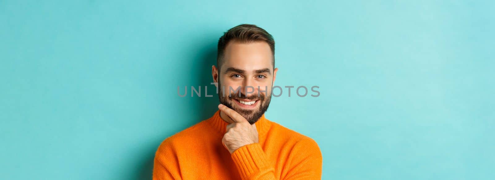 Close-up of handsome and confident man with beard smiling, looking thoughtful at camera, have plan, standing over light blue background.