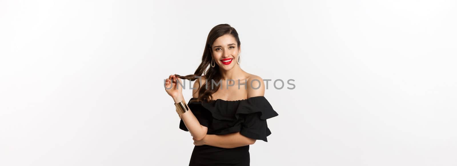 Beauty and fashion concept. Flirty woman in black dress, red lips, playing with hair and listening, looking at camera with interest, standing over white background by Benzoix