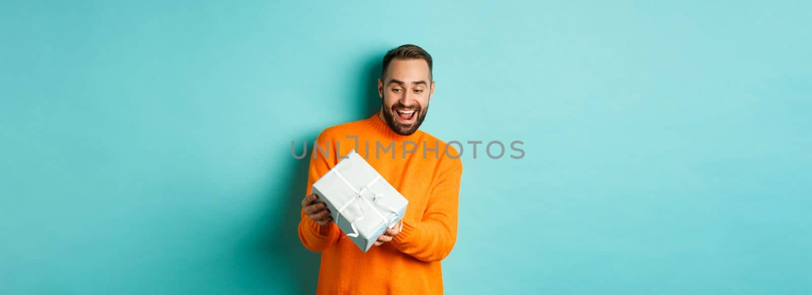 Holidays and celebration concept. Excited man receiving gift, looking happy at present and smiling, standing over blue background.