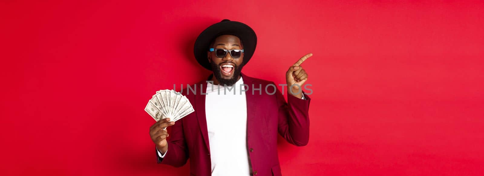 Handsome and stylish Black man pointing fingers left while showing money, holding dollars and demonstrating logo, standing over red background.