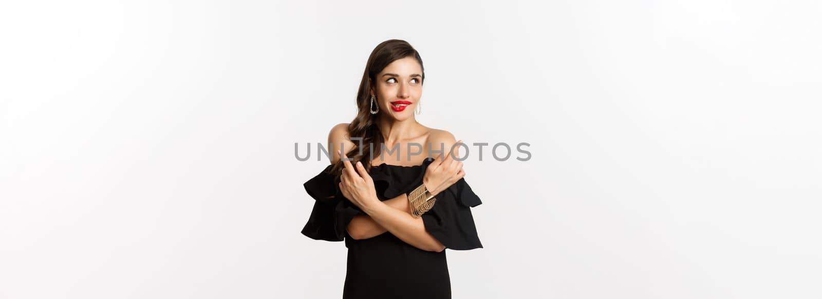 Fashion and beauty. Beautiful glamour woman in black dress, making choice, biting lip from temptation and pointing sideways, standing over white background.