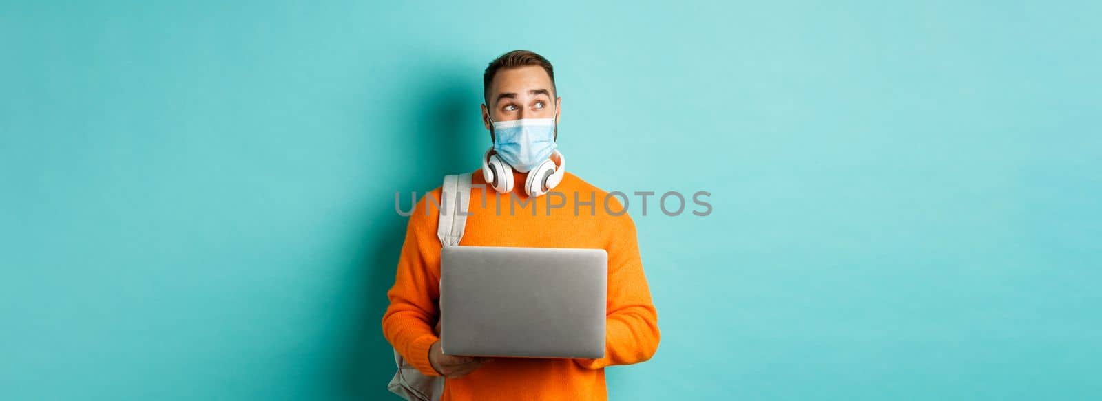 Happy young freelancer in medical mask, working on laptop, looking aside thoughtful, standing over turquoise background.