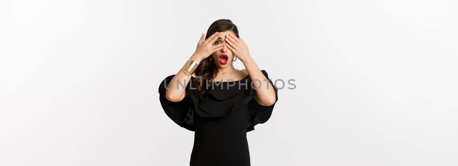 Fashion and beauty. Image of shocked glamour woman in black dress, covering eyes but peeking through fingers startled, standing over white background by Benzoix