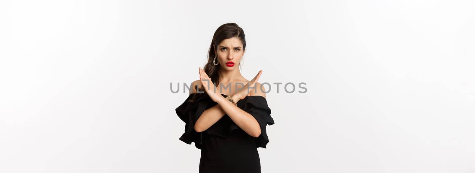 Fashion and beauty. Serious and confident female model in black dress, showing cross sign and frowning, stop gesture, telling no, standing over white background by Benzoix