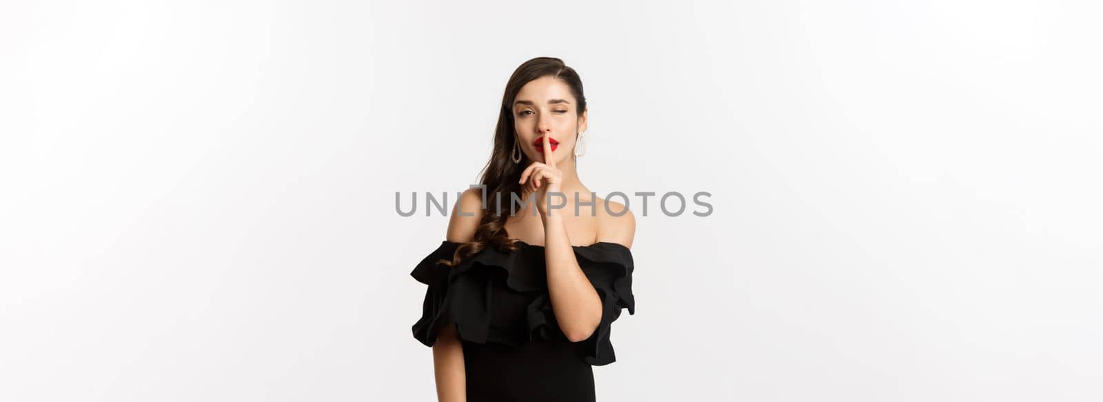 Fashion and beauty. Coquettish young woman in black dress, red lips, winking at camera and making hush sign, standing over white background.