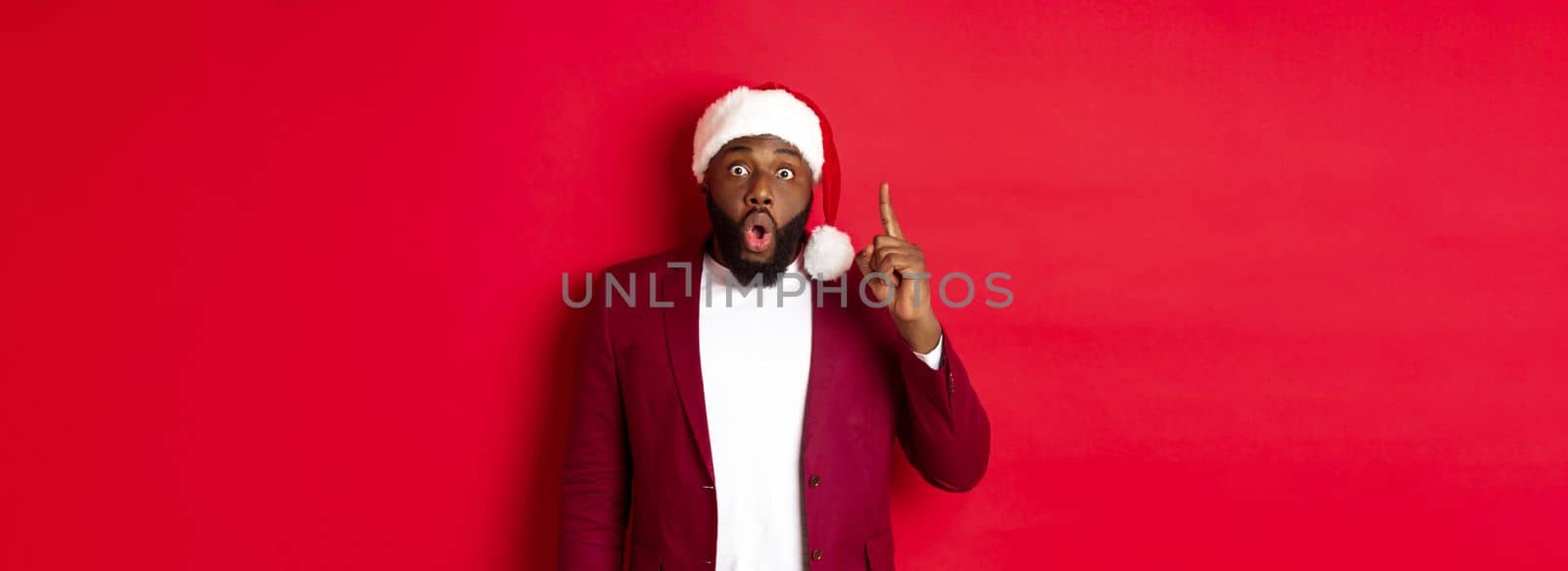 Christmas, party and holidays concept. Excited Black man in santa hat having an idea, raising finger up, suggesting plan, standing against red background by Benzoix