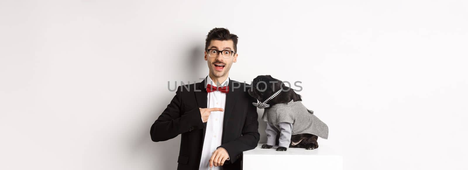 Cheerful guy standing with his cute pug, pointing finger at dog wearing party costume, posing over white background.