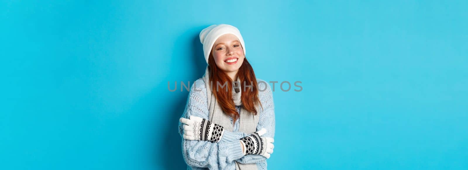 Winter and holidays concept. Smiling redhead girl in beanie, gloves and sweater getting warm after going outside, standing over blue background by Benzoix