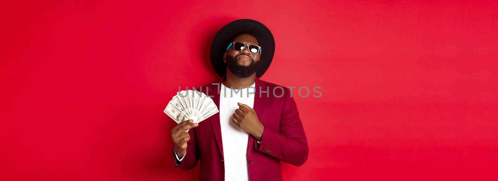 Sassy and cool african american man in sunglasses and hat, pointing at himself and showing dollars, looking confident against red background.