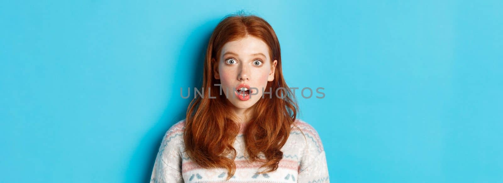 Close-up of shocked redhead girl drop jaw in awe, staring with amazement at camera, standing against blue background.