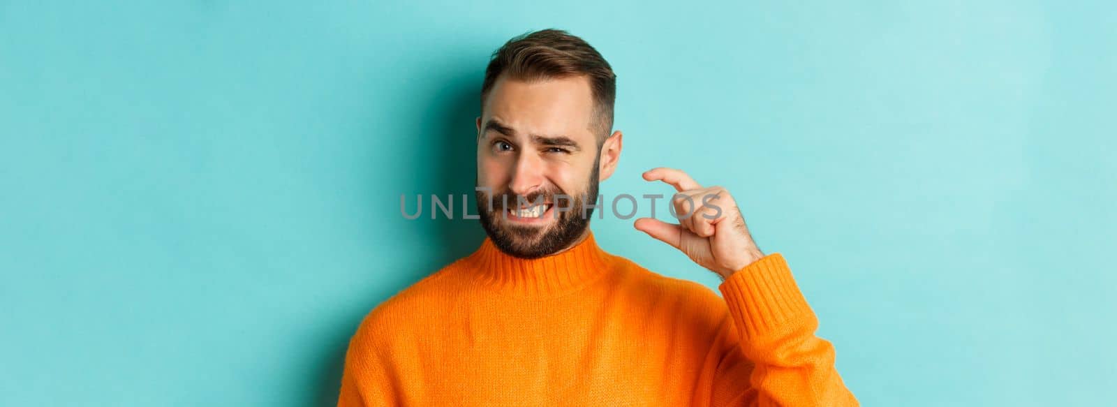 Close-up of awkward man showing small or little with fingers, standing displeased over light blue background by Benzoix