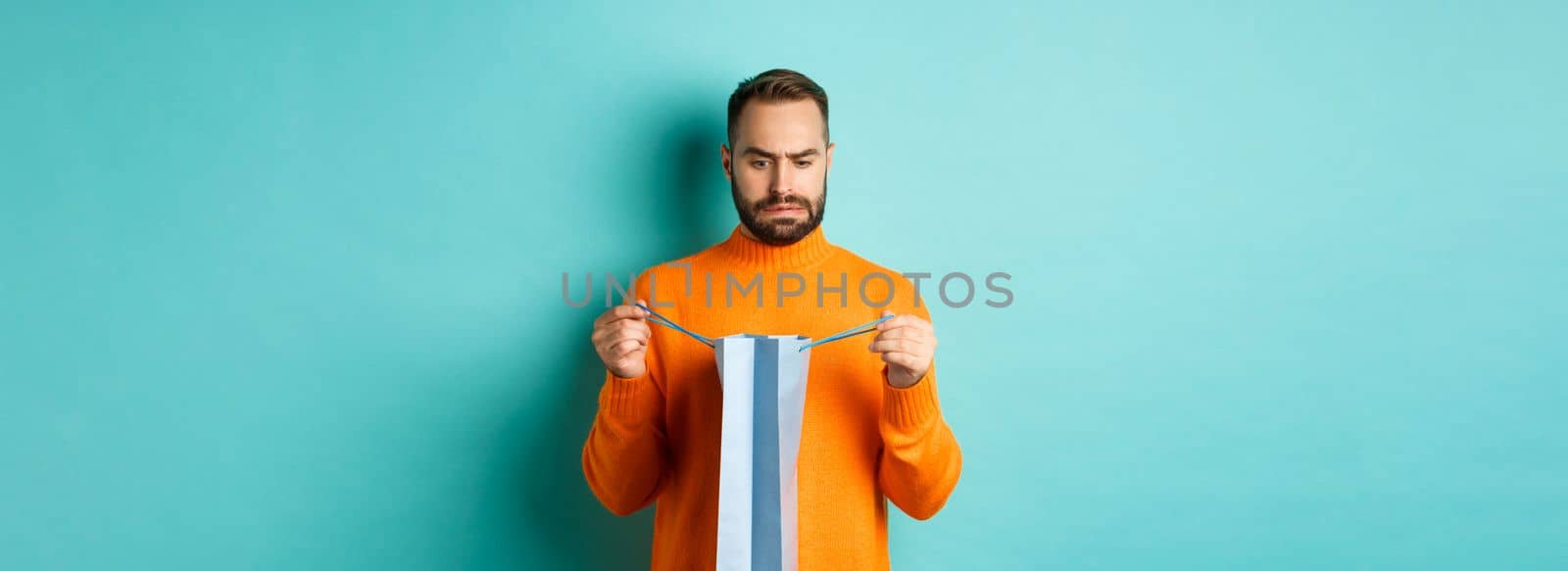 Disappointed man open shopping bag and dislike gift, frowning displeased, standing in orange sweater against turquoise background.