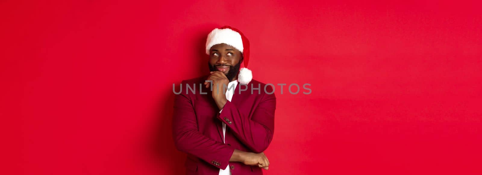 Christmas, party and holidays concept. Smiling african american man making new year plans, looking thoughtful at upper left corner, wearing santa hat, red background.