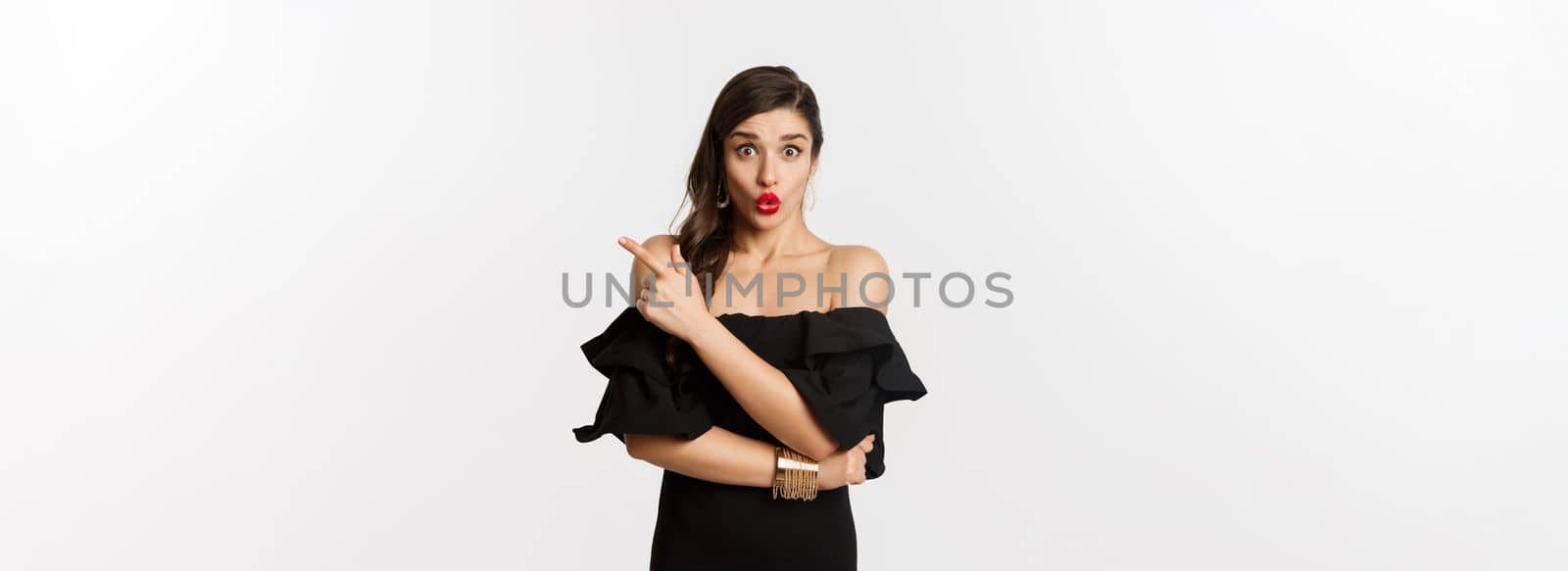 Fashion and beauty. Surprised woman in black dress pointing finger at upper right corner, showing advertisement, standing over white background.