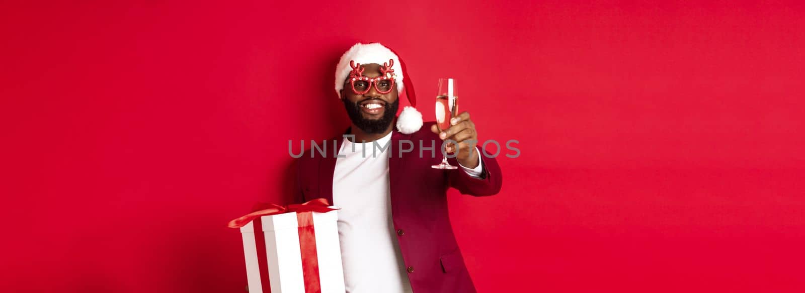 Christmas. Handsome african american man in party glasses and santa hat, holding new year gift and glass of champagne, wishing happy holidays, red background.