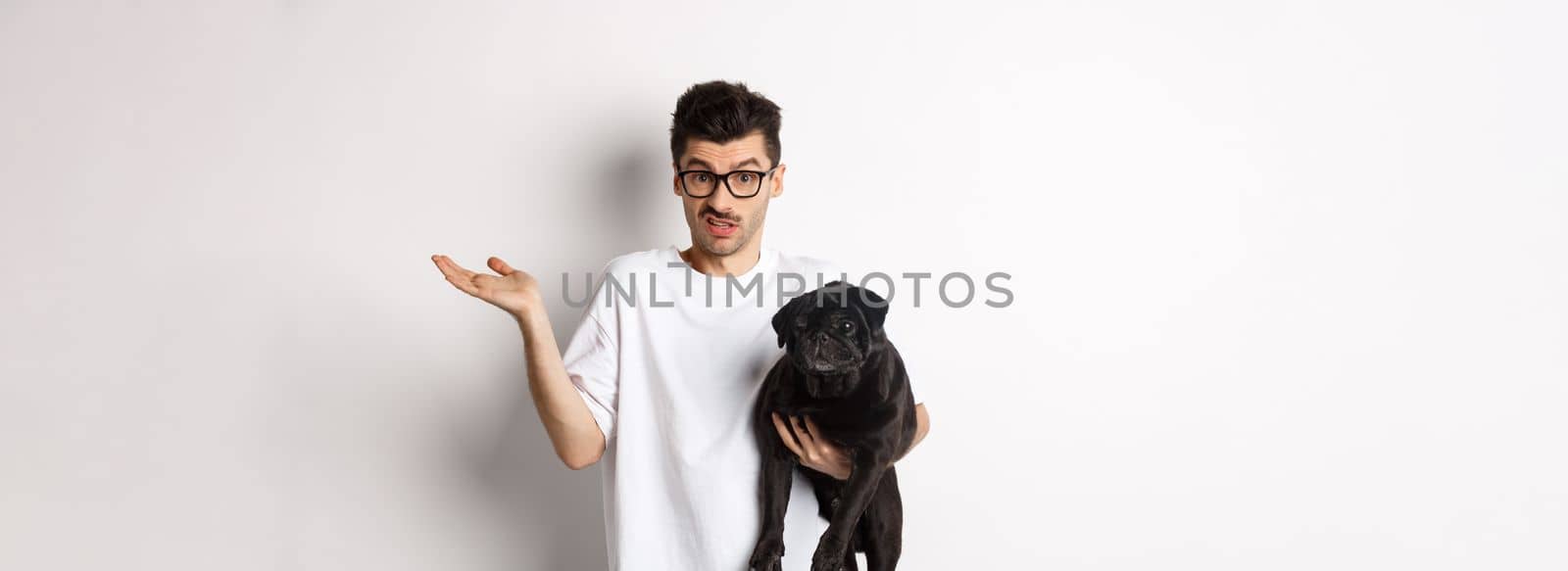 Image of handsome young man holding black pug and looking confused. Guy shrugging shoulders and staring indecisive at camera, carry dog in arm, white background.