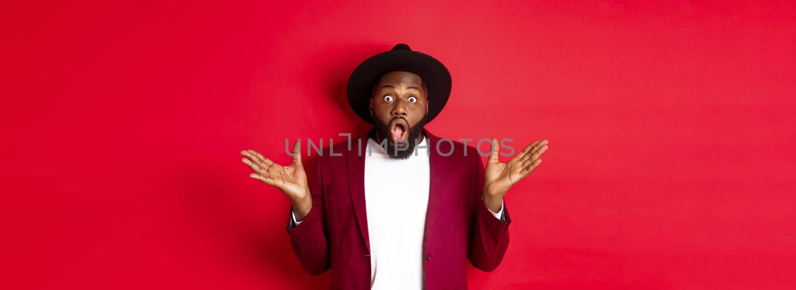 Fashion and party concept. Impressed Black man in classy outfit staring with complete disbelief at camera, gasping and looking surprised, standing over red background.