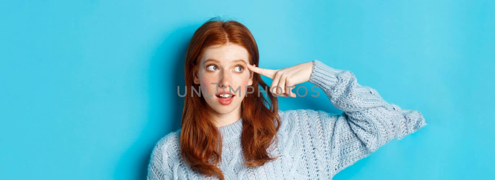 Close-up of cute redhead girl searching for solution, pointing at head and looking left at logo, standing over blue background.