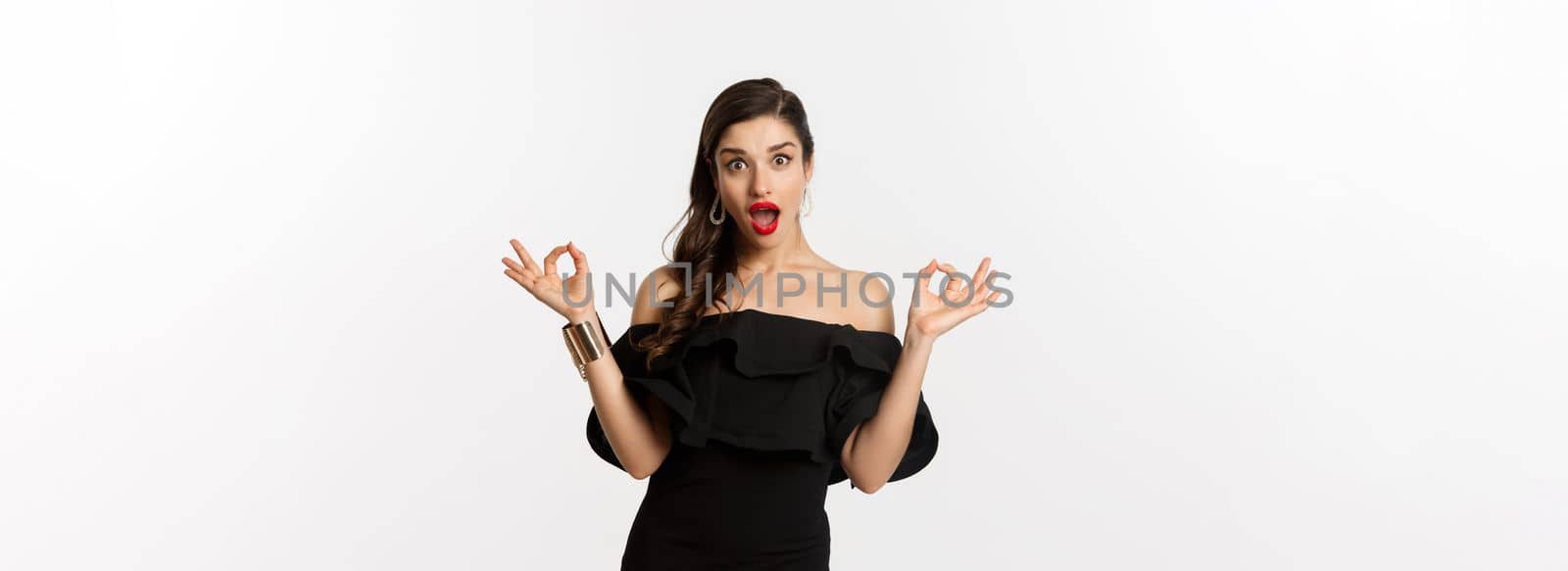 Fashion and beauty. Attractive brunette woman in black dress, showing okay signs and staring excited, approve and recommend, standing over white background.